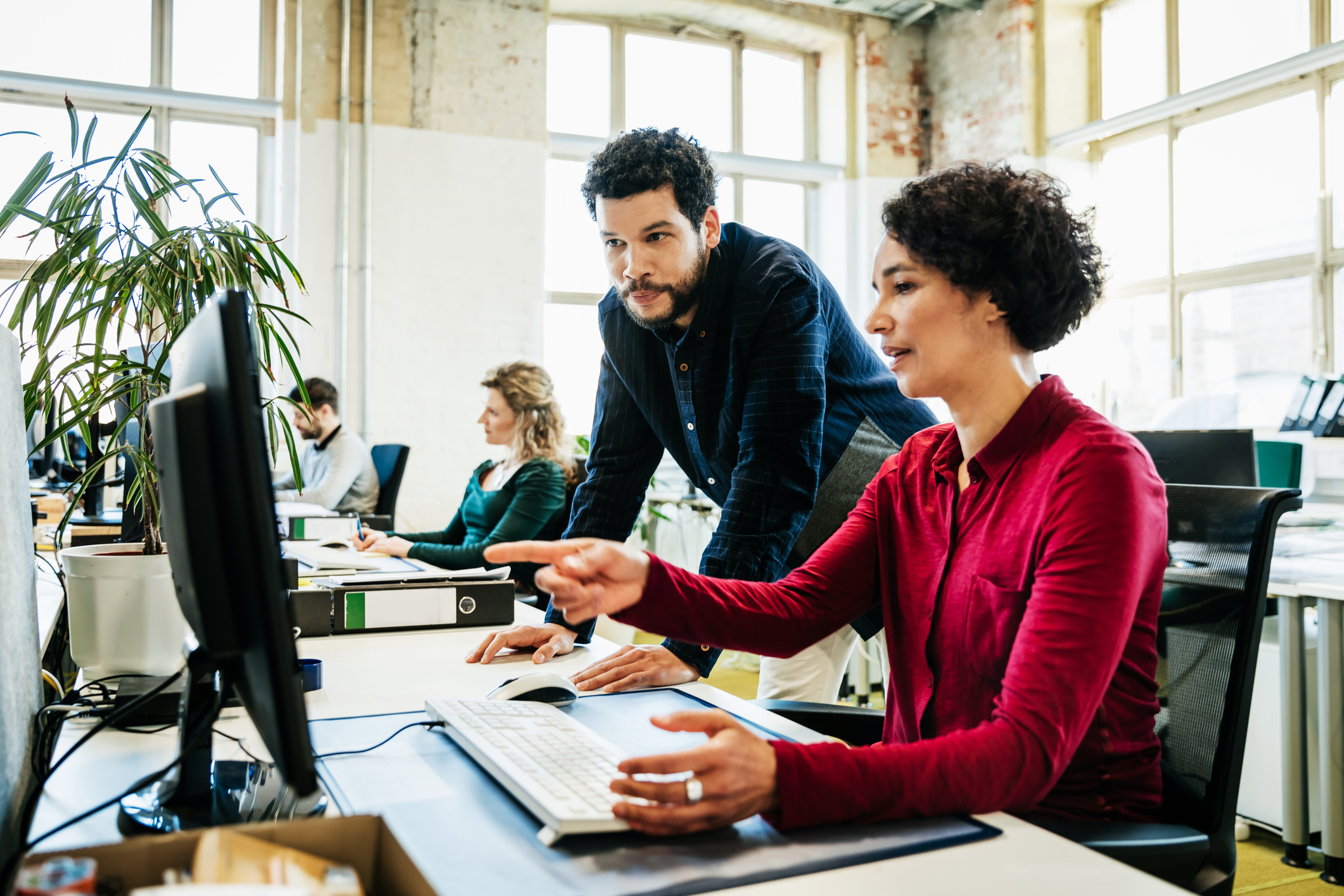 two colleagues in an office at the same desk. One sat pointing at the computer screen in front of her, the other male colleague standing with hands on the desk, looking at the screen.